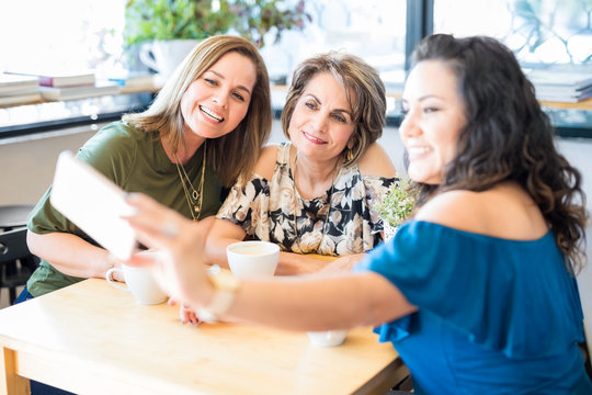 A group of three middle aged women are gathered together in front of a phone for a selfie at a cafe